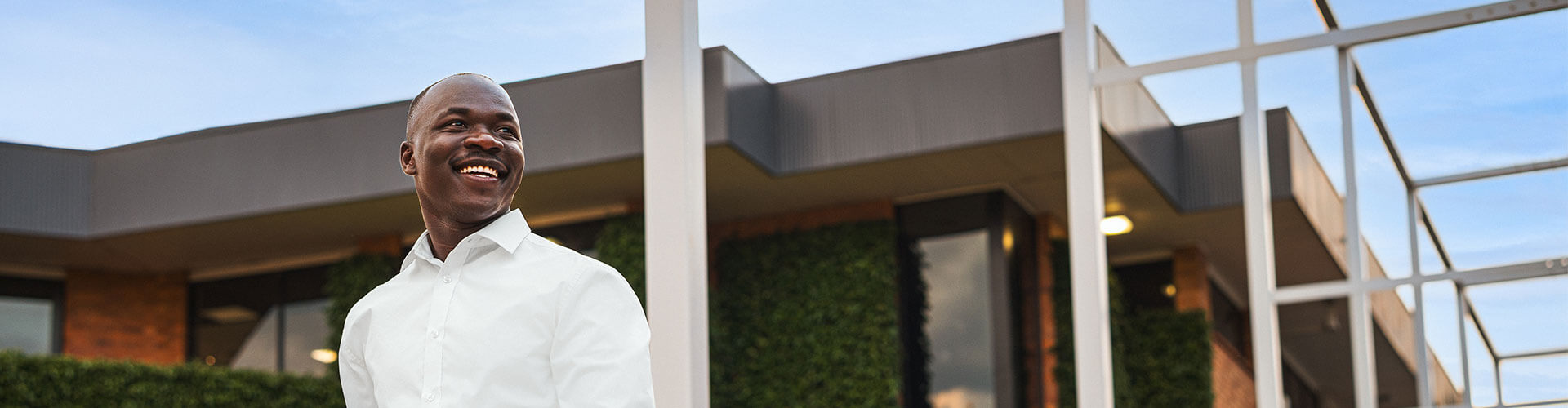 A man in a white shirt stands outside a modern building with large windows and greenery, smiling and looking to his left.
