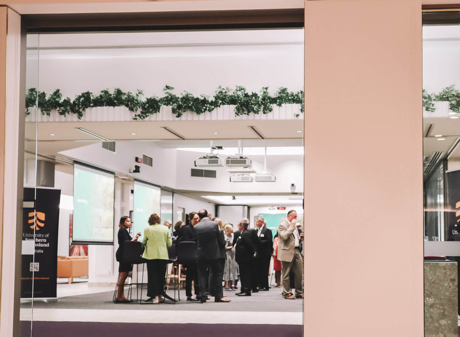 A group of people in business attire are conversing and networking in a modern conference room with large screens and plants on the ledge above.