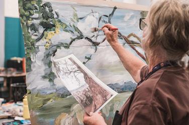 Person painting a landscape on a canvas while holding a reference photo of trees; paint bottles and art supplies are on the table nearby.
