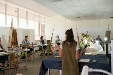 A woman stands at a table addressing a group of people seated at desks in an art classroom with easels, flowers, and bright daylight.