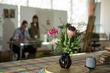 A black vase with pink and purple flowers sits on a table; two blurred people are in the background near a whiteboard.