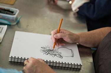 A person sketches a flower in a vase on a spiral-bound notebook using a pencil; art supplies are nearby on a metallic table.