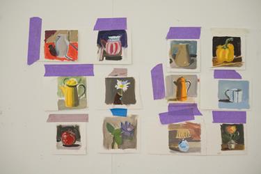 Twelve small still life paintings, mostly of pitchers, fruit, and flowers, are taped to a white wall with pieces of purple and brown tape.