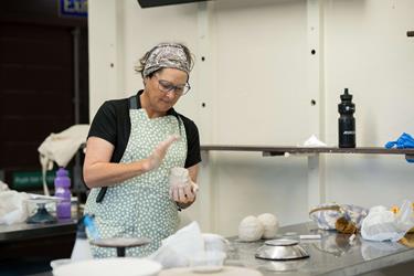 A person wearing an apron and headband shapes clay with their hands at a worktable. Various tools and materials are scattered on the table.