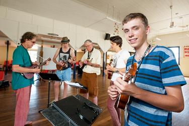 A group of people stand in a room playing ukuleles, with a young man in a striped shirt smiling at the camera in the foreground.