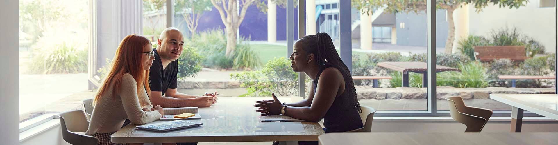 Three people sit at a table in a bright room with large windows, engaged in conversation with notebooks and pens in front of them.