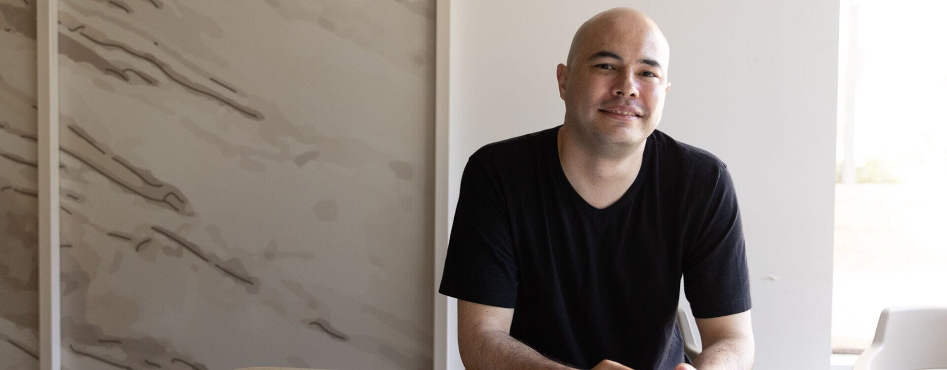 A man with a shaved head wearing a black t-shirt sits at a table, smiling gently, in a well-lit room with a neutral-toned wall in the background.