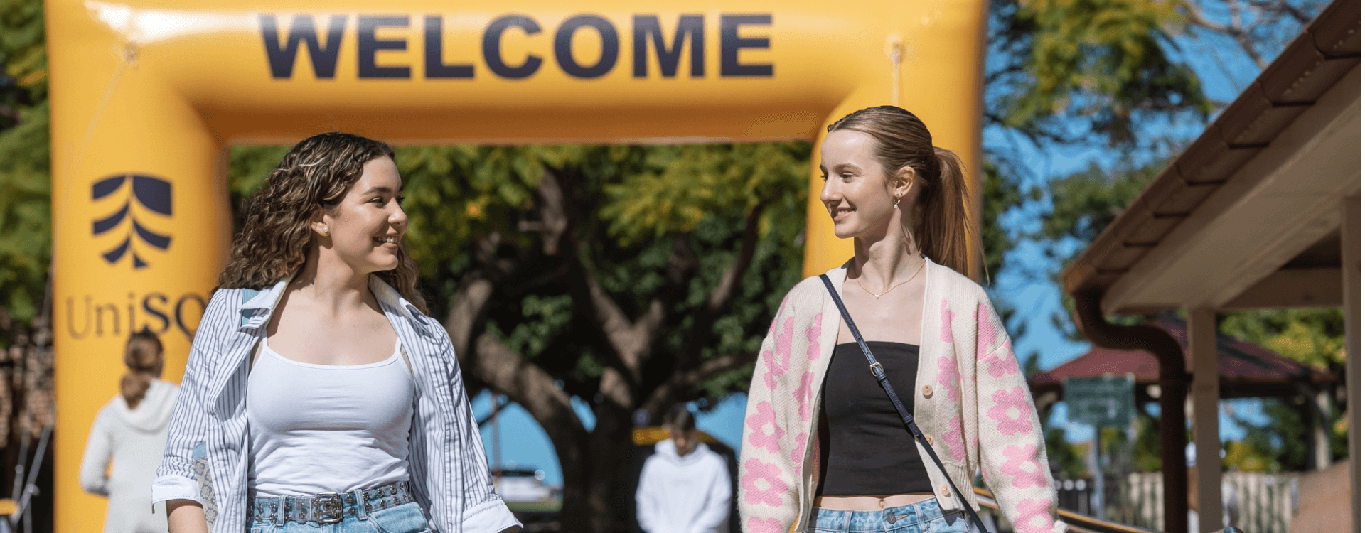 Two young women walk and smile at each other under a large yellow "WELCOME" arch outdoors, with people and trees in the background.