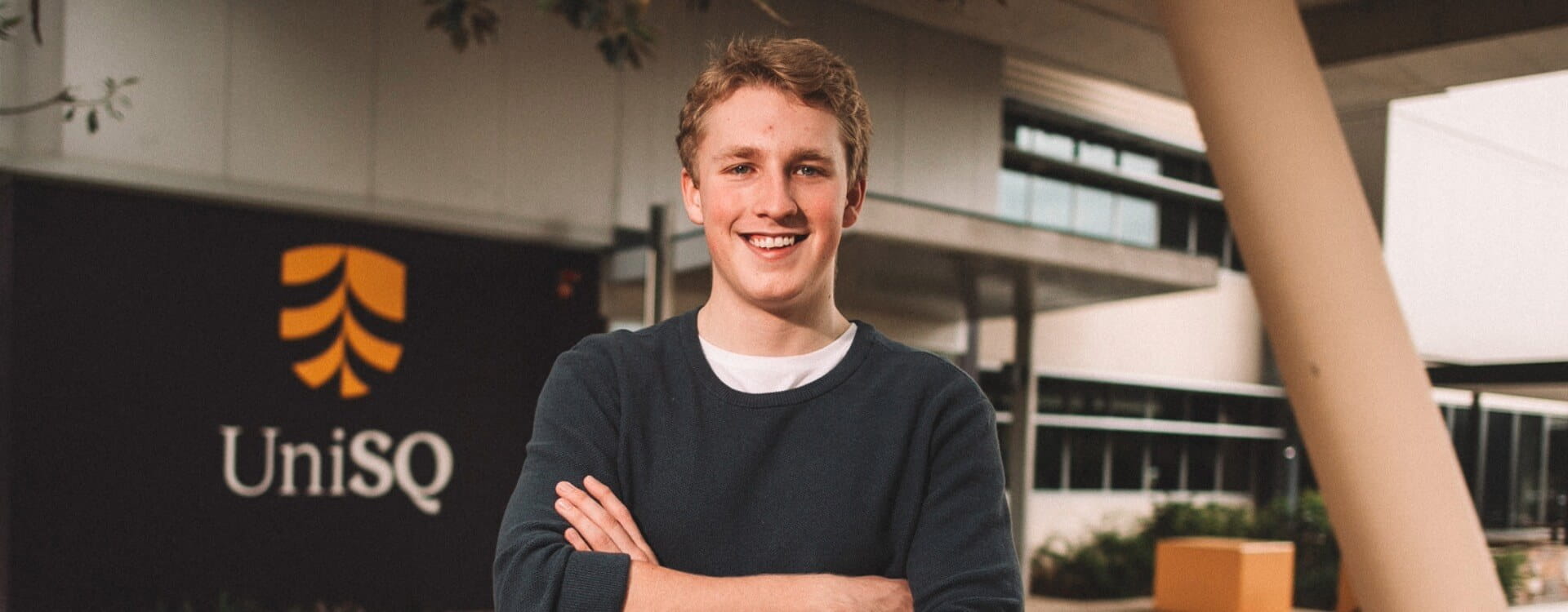 A young man with arms crossed stands outside a building with a UniSQ sign visible in the background.