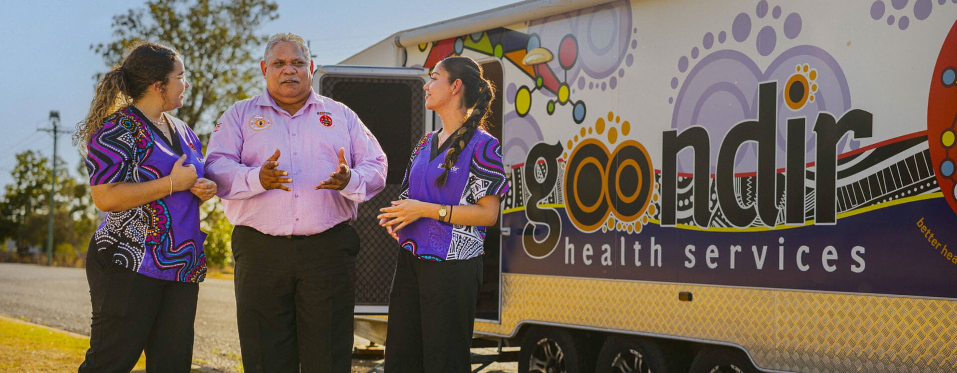 Three people stand and talk in front of a Goondir Health Services mobile clinic decorated with colorful Indigenous artwork.