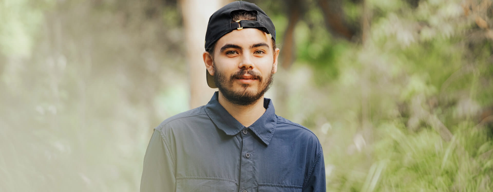 A man wearing a navy shirt and a backward black cap stands outdoors with greenery in the background.