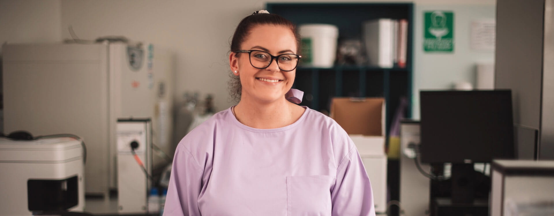Person wearing glasses and a lavender top smiles in a laboratory setting with various equipment in the background.