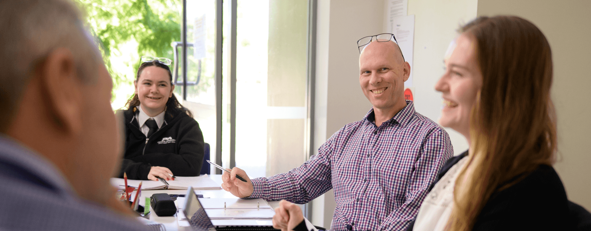 Four people sit around a table in an office, engaged in discussion. Two people are facing the camera, smiling, with papers and laptops on the table.