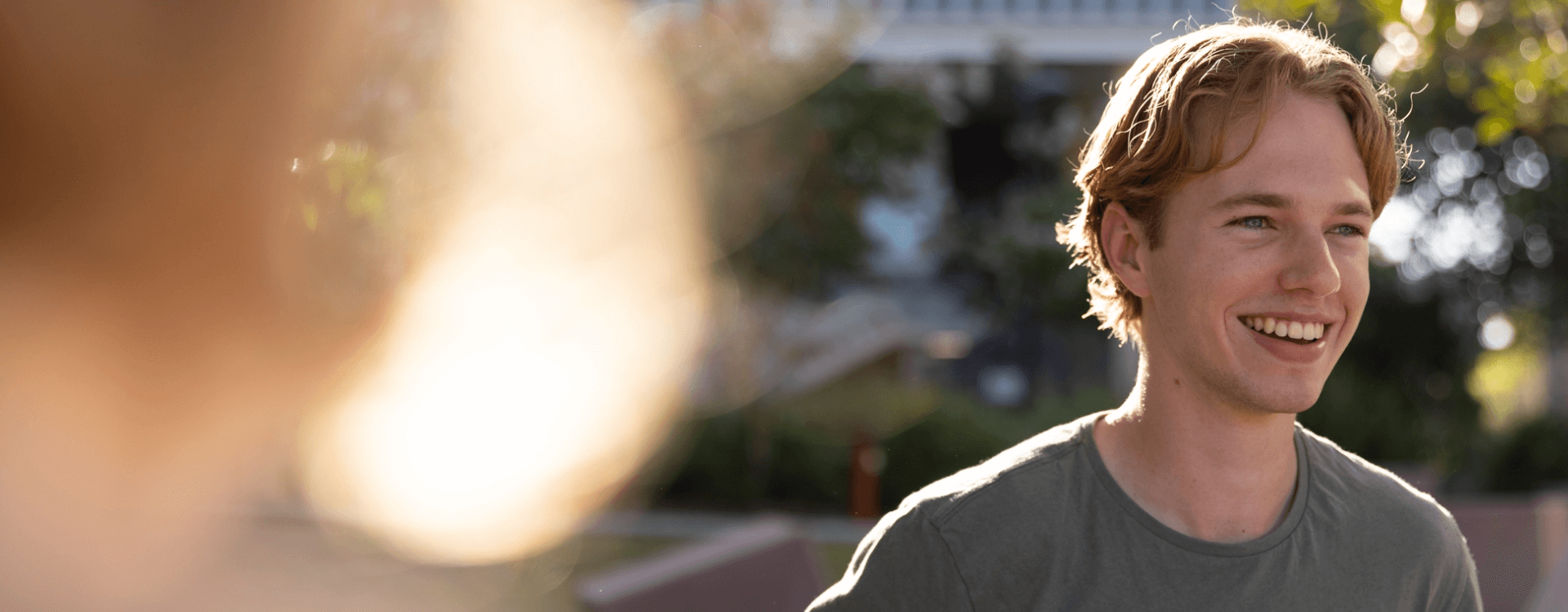 A young man with blond hair and a gray shirt smiles outdoors in natural sunlight, with blurred greenery and another out-of-focus person in the foreground.