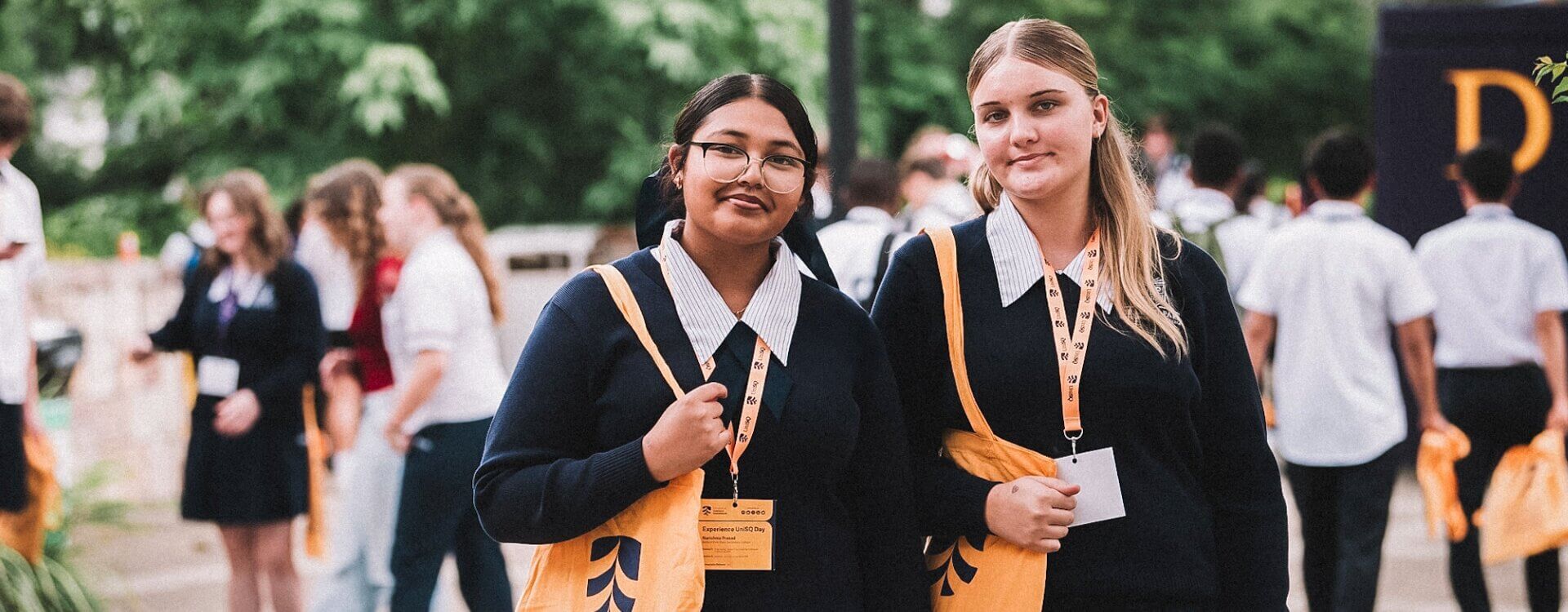 Two young women in navy uniforms with yellow tote bags stand outdoors among a group of people, wearing name badges and looking at the camera.