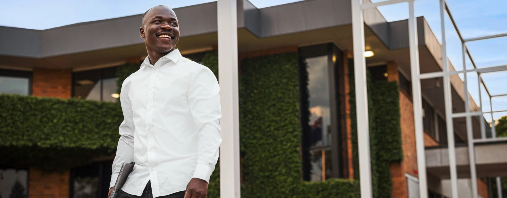 A man in a white shirt holds a tablet and smiles while standing outside a modern building with greenery on the walls.