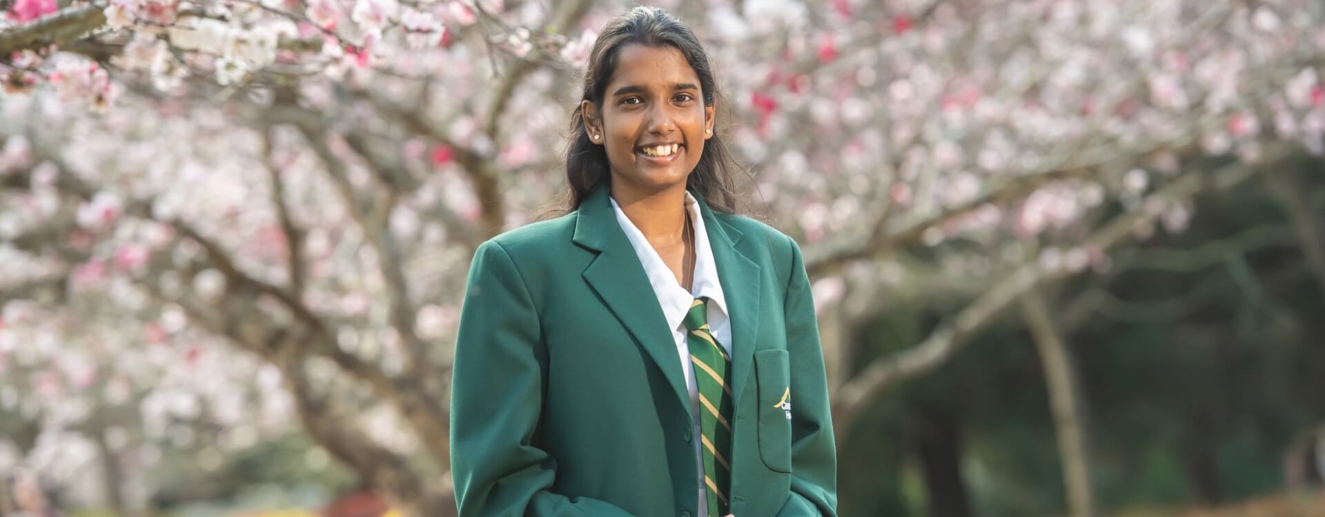 A student in a green school blazer and striped tie stands outdoors in front of blossoming trees, smiling at the camera.
