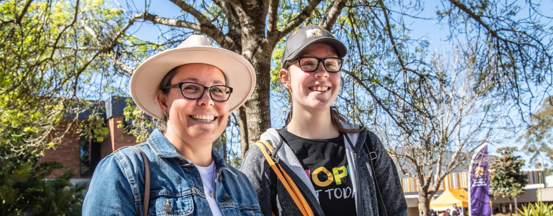 Two people wearing glasses and hats smile outdoors, standing in front of a tree with buildings and other people visible in the background.