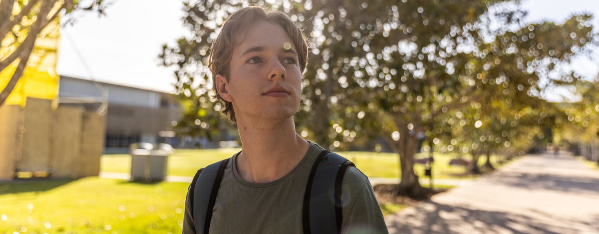A young person wearing a backpack stands outdoors on a sunny day, with trees and a walkway in the background.