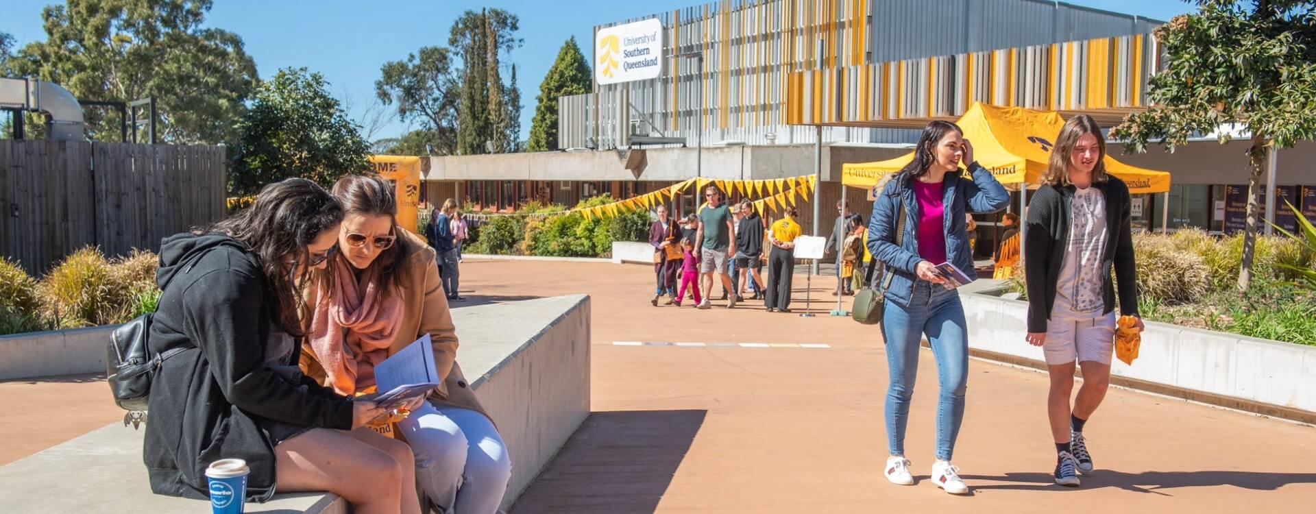 Students walk and sit on a modern university campus, with buildings and trees in the background on a sunny day.