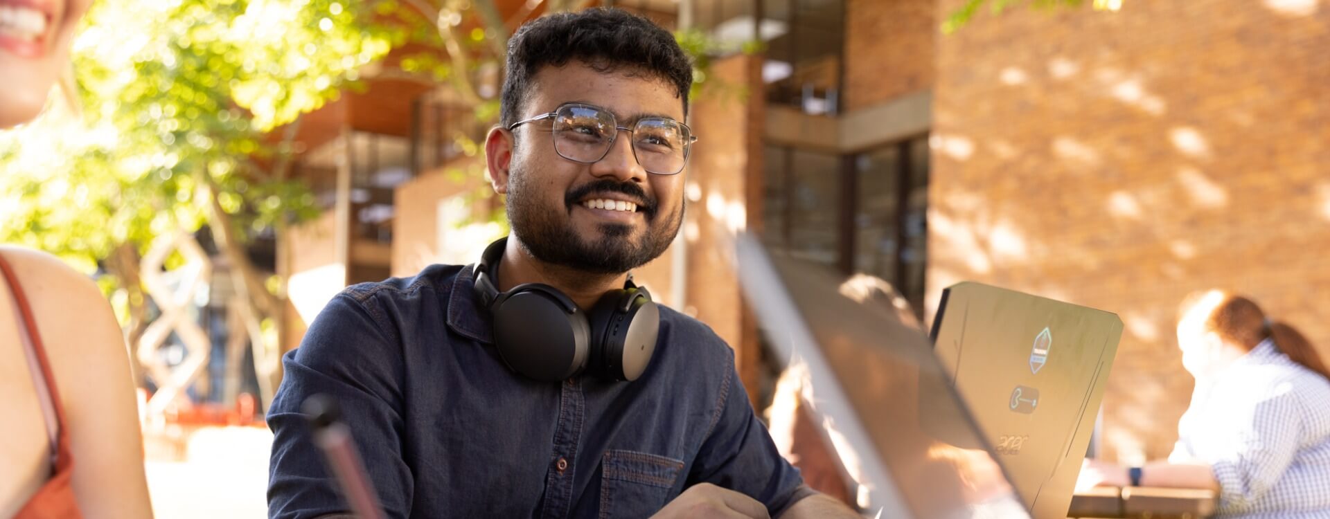 A man wearing glasses and headphones around his neck smiles while sitting outdoors at a table with a laptop. Other people are visible in the background.
