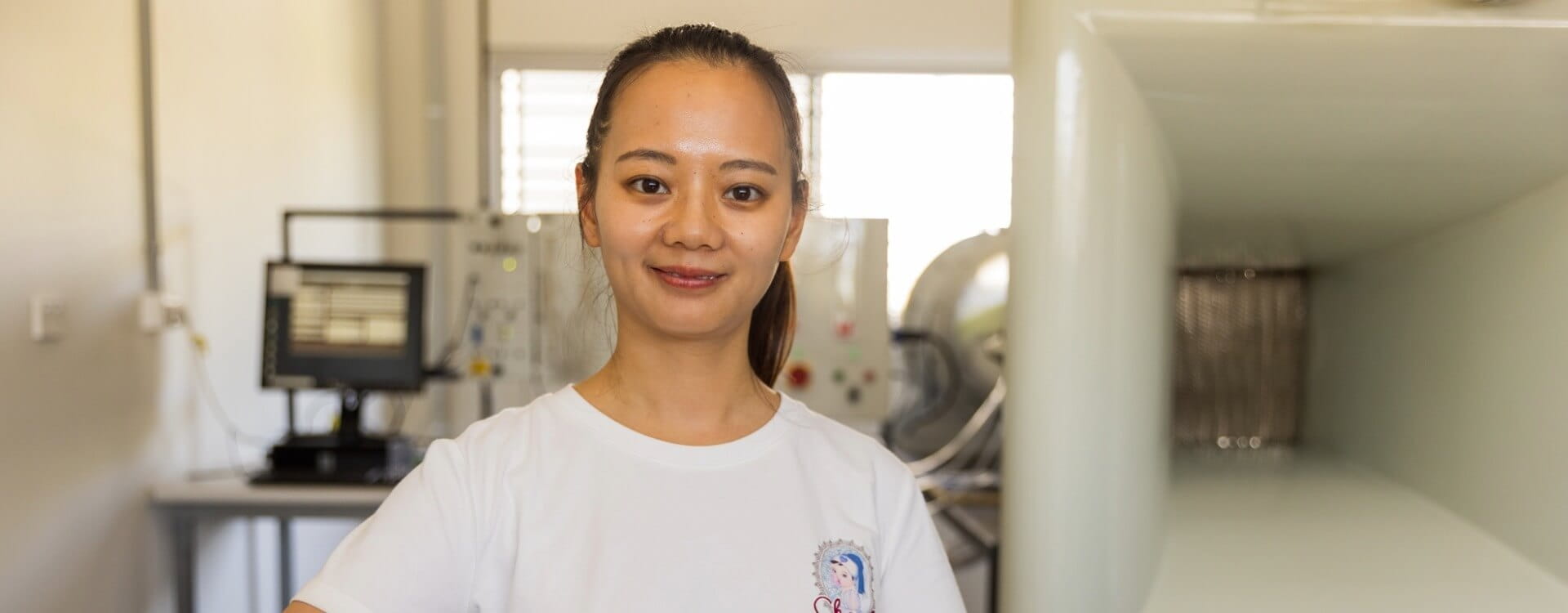 A woman stands in a laboratory, wearing a white t-shirt, with scientific equipment and a computer visible in the background.