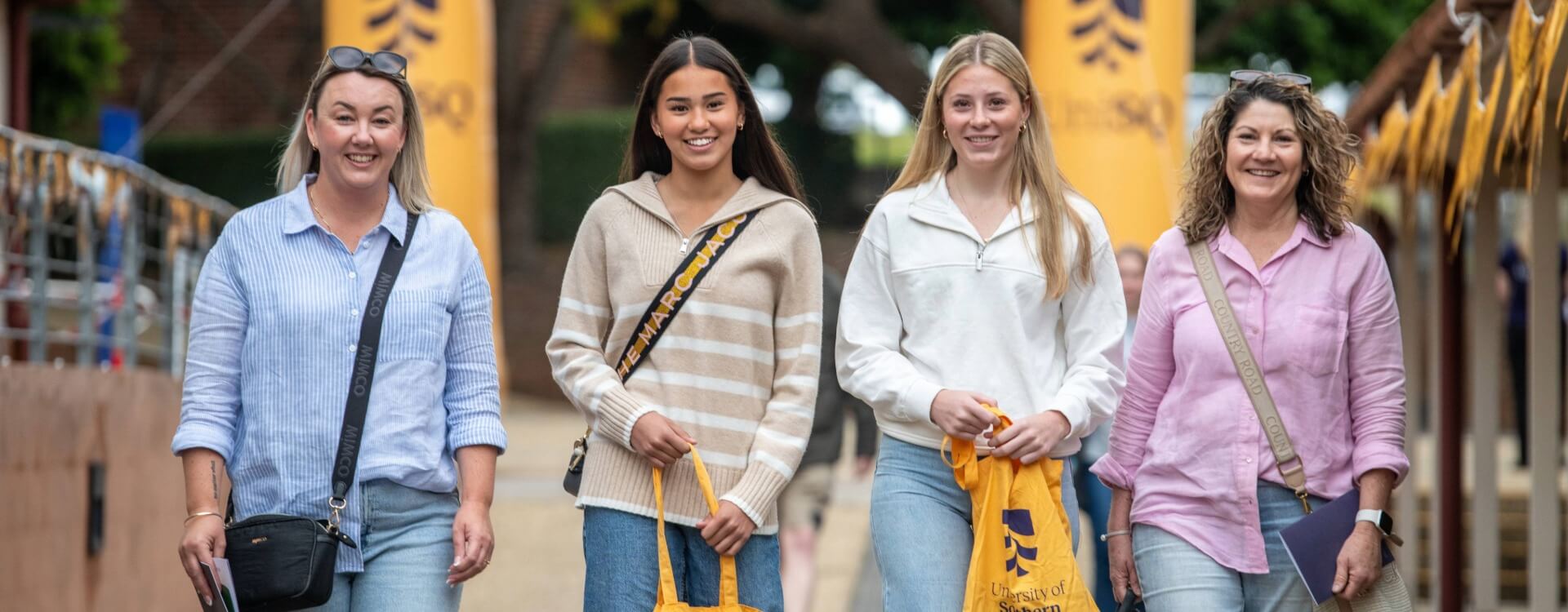 Four women walking outside on a sidewalk, holding yellow bags, with university banners and trees in the background.
