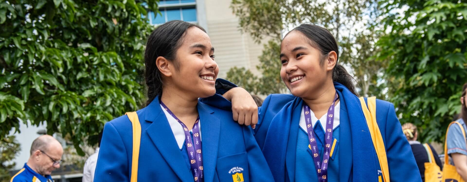 Two students in blue blazers and lanyards smile and walk outdoors, with one student’s arm around the other’s shoulder. Trees and people are in the background.