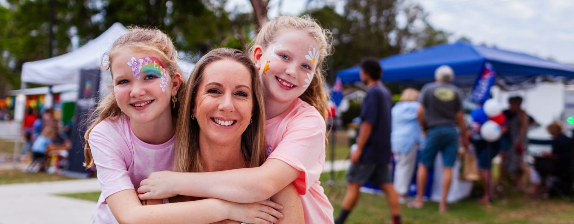 A woman smiles while two children with face paint hug her outdoors at a community event with tents, balloons, and people in the background.
