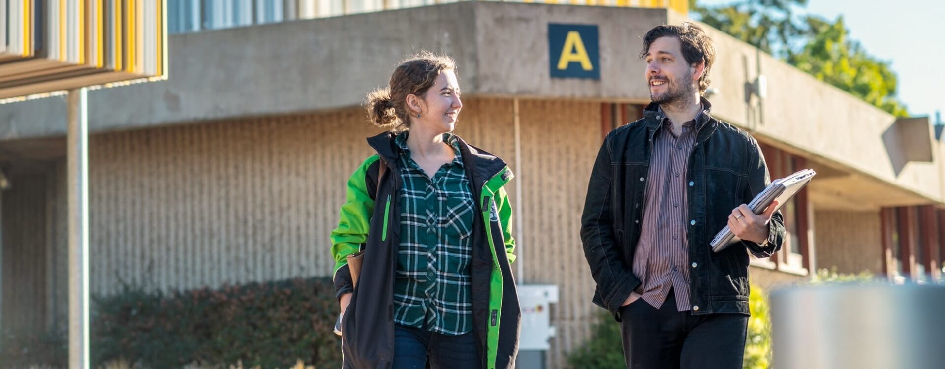 Two people walk outdoors near a building marked with a large letter “A”; one carries folders and papers, both are smiling and casually dressed.