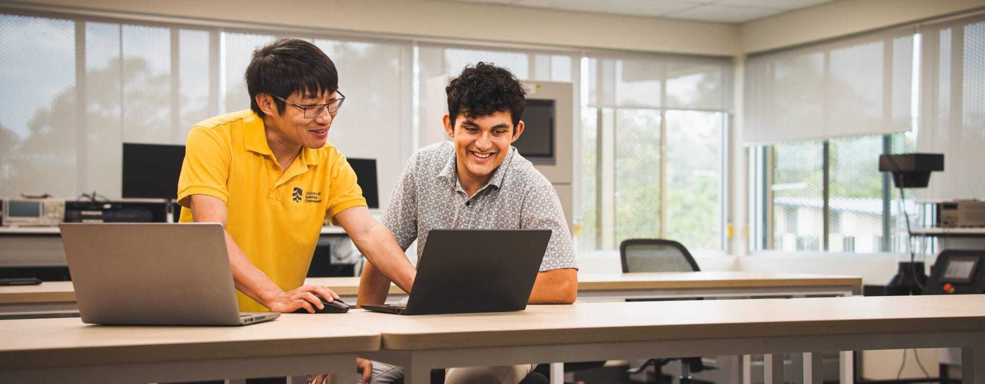 Two men in a bright classroom look at a laptop together, one standing and pointing at the screen while the other sits and smiles.