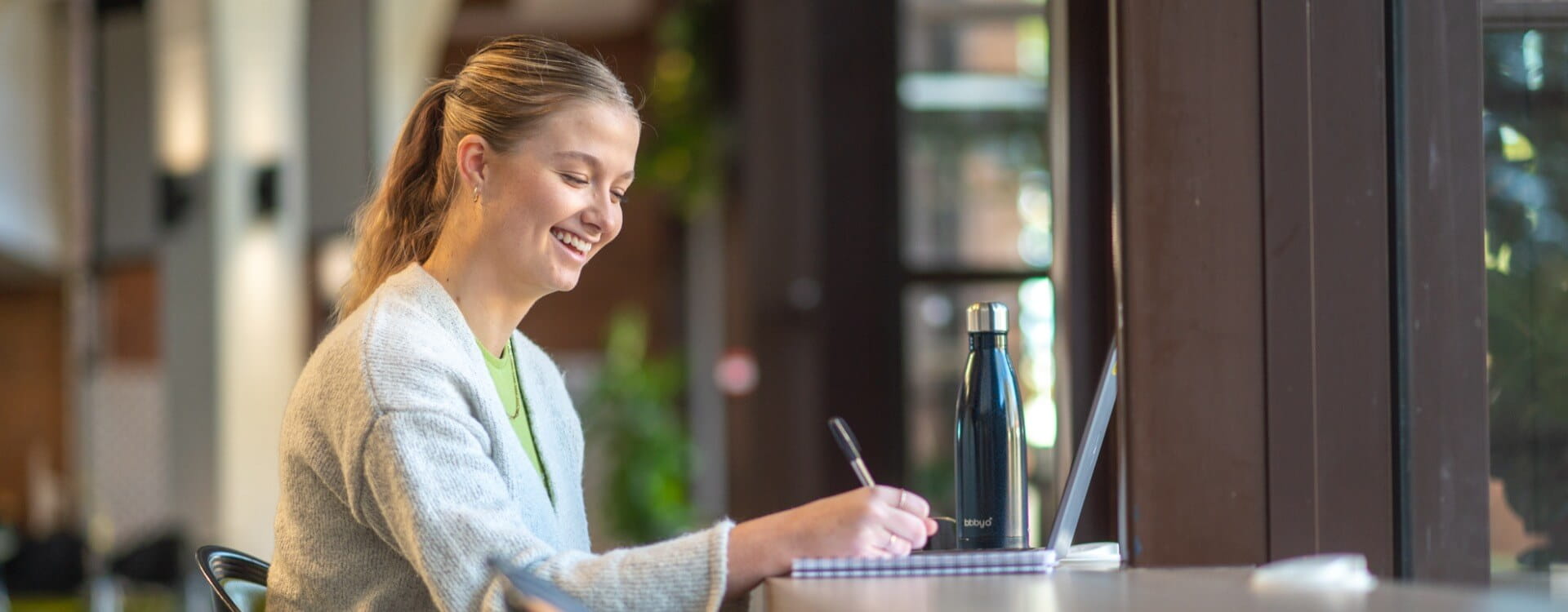 A person sits at a table writing in a notebook with a laptop and a water bottle nearby, smiling and looking at the screen.