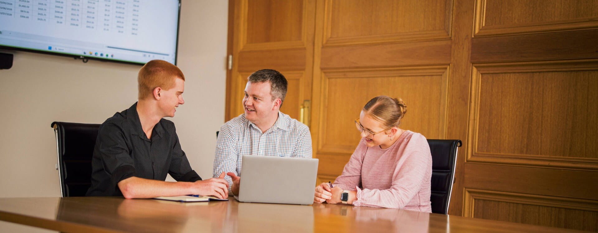 Three people sit at a conference table with a laptop, having a discussion. A screen with data is visible on the wall behind them.