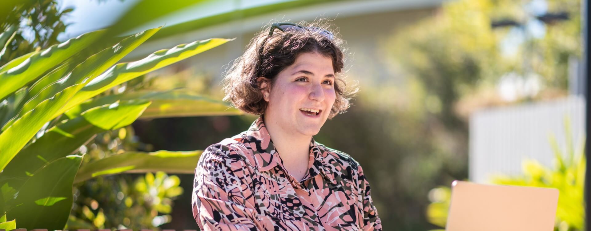 A person with short brown hair sits outdoors, wearing a patterned shirt and smiling while using a laptop. Large green leaves are visible in the foreground.