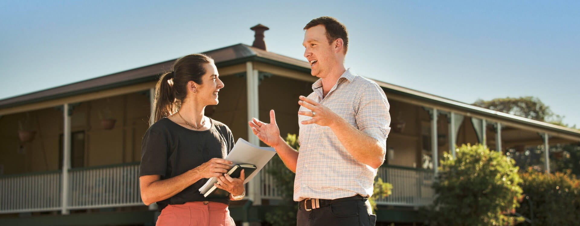 Two people stand outdoors in front of a house, engaged in conversation. One person holds a tablet and notepad while the other gestures with their hands.