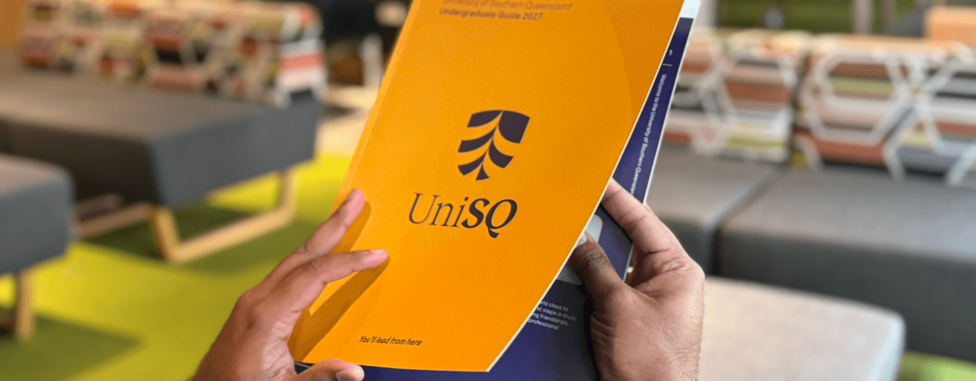 A person holds a University of Southern Queensland (UniSQ) undergraduate guide booklet in a lounge area with sofas and bookshelves in the background.