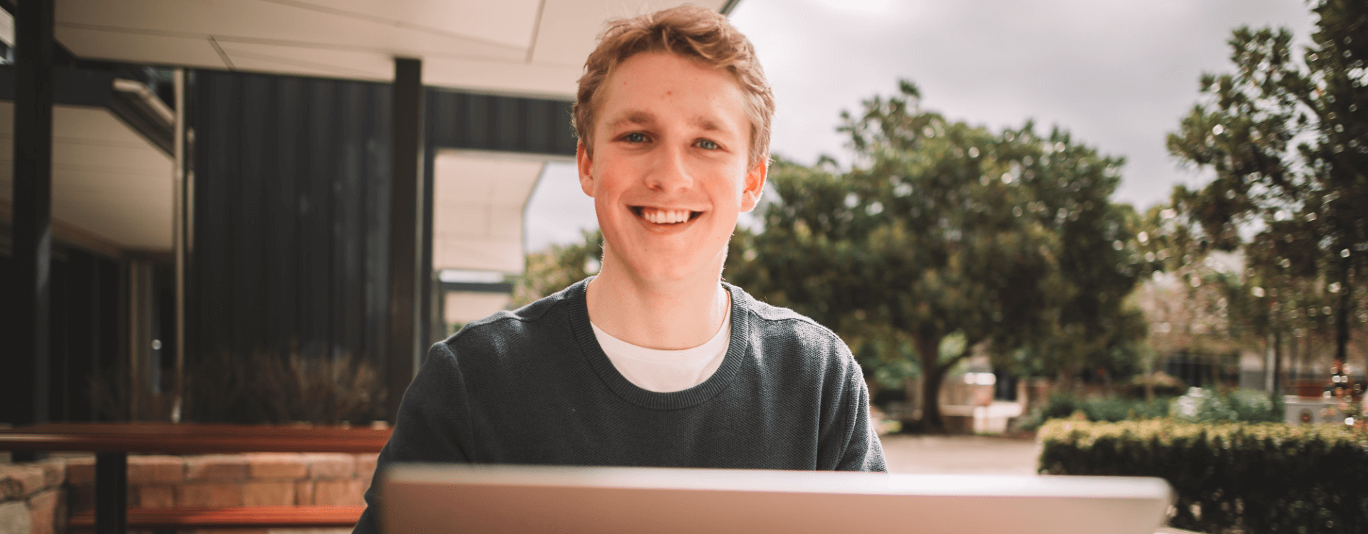 A young man sitting outdoors in front of a laptop, smiling at the camera, with trees and a modern building in the background.