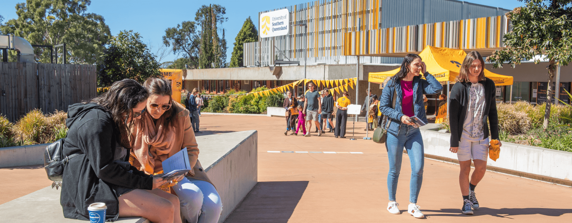 Students walk and sit outdoors on a university campus, with buildings, yellow tents, and trees visible in the background on a sunny day.