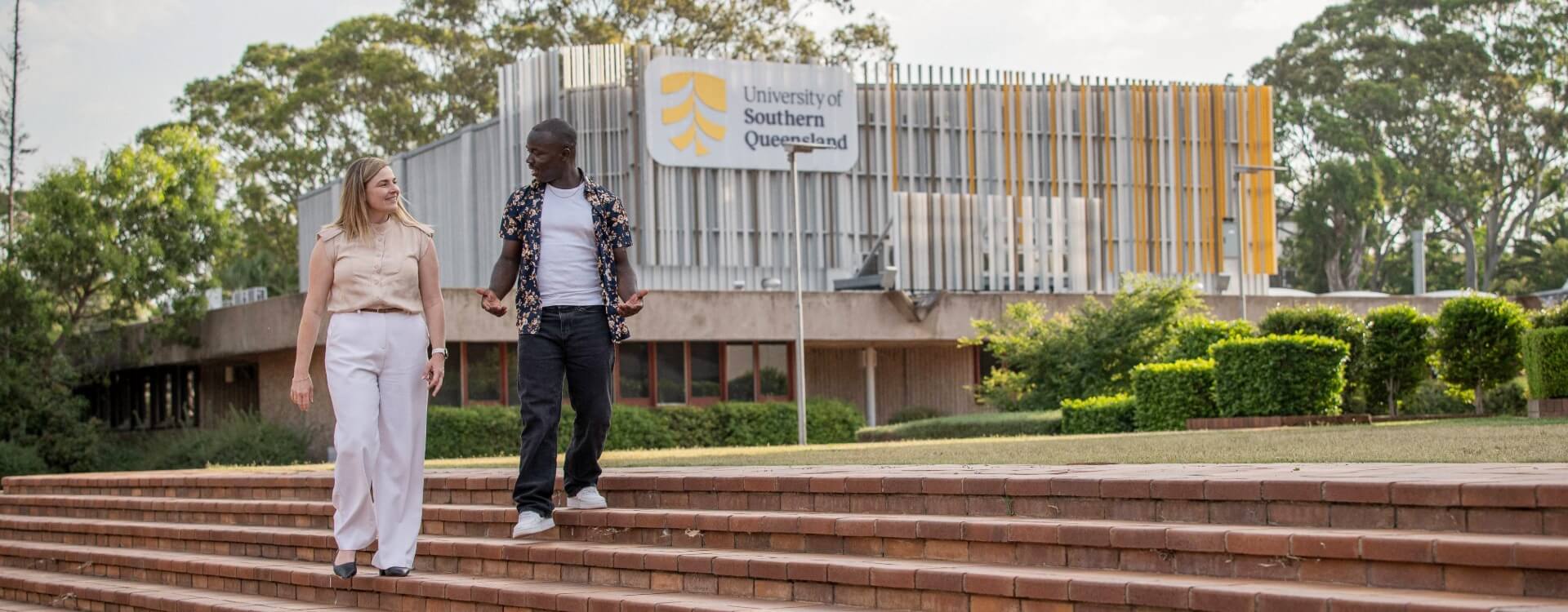 Two people walk down steps outside the University of Southern Queensland building on a clear day, with trees and greenery in the background.
