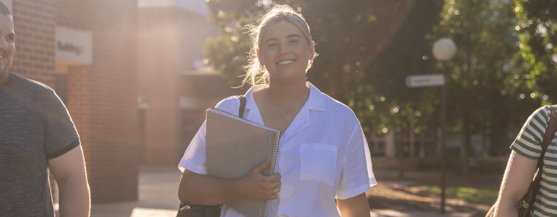 Person in a white shirt holding notebooks and smiling while walking outdoors, with sunlight in the background and two others partially visible beside them.