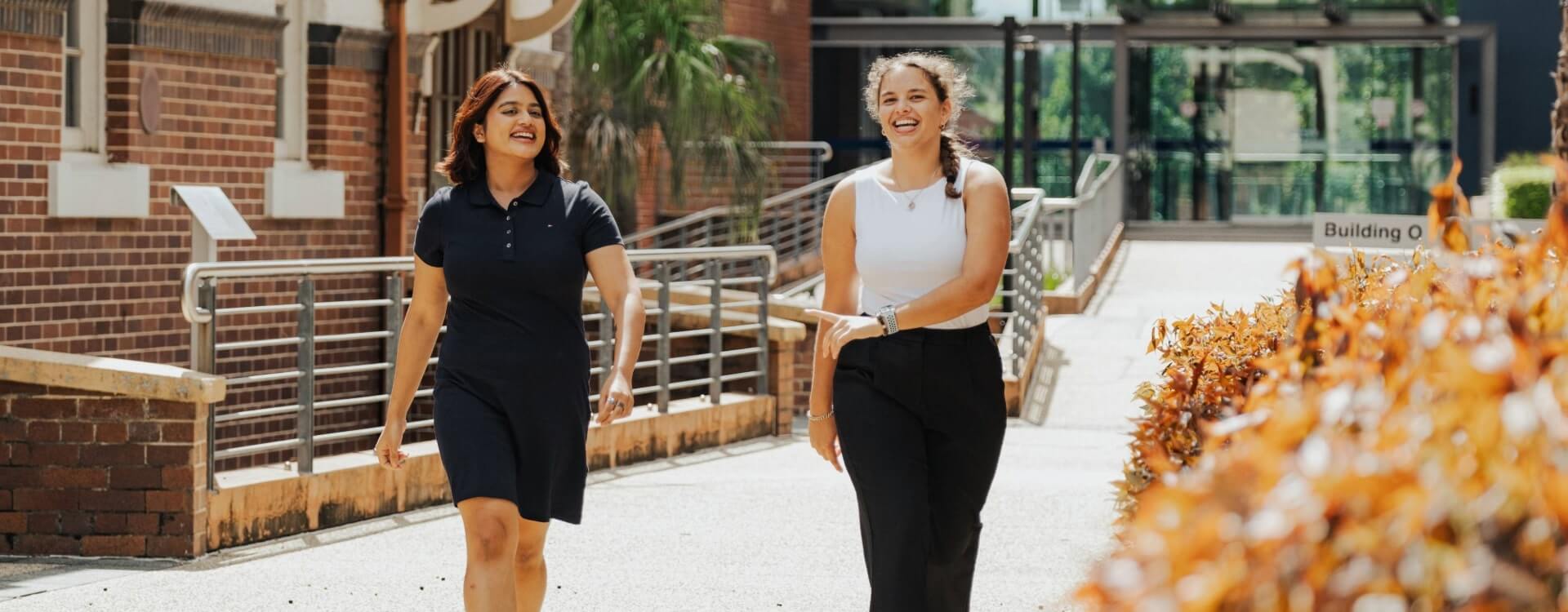 Two women are walking and talking outside on a sunny day, near a brick building and landscaped bushes.