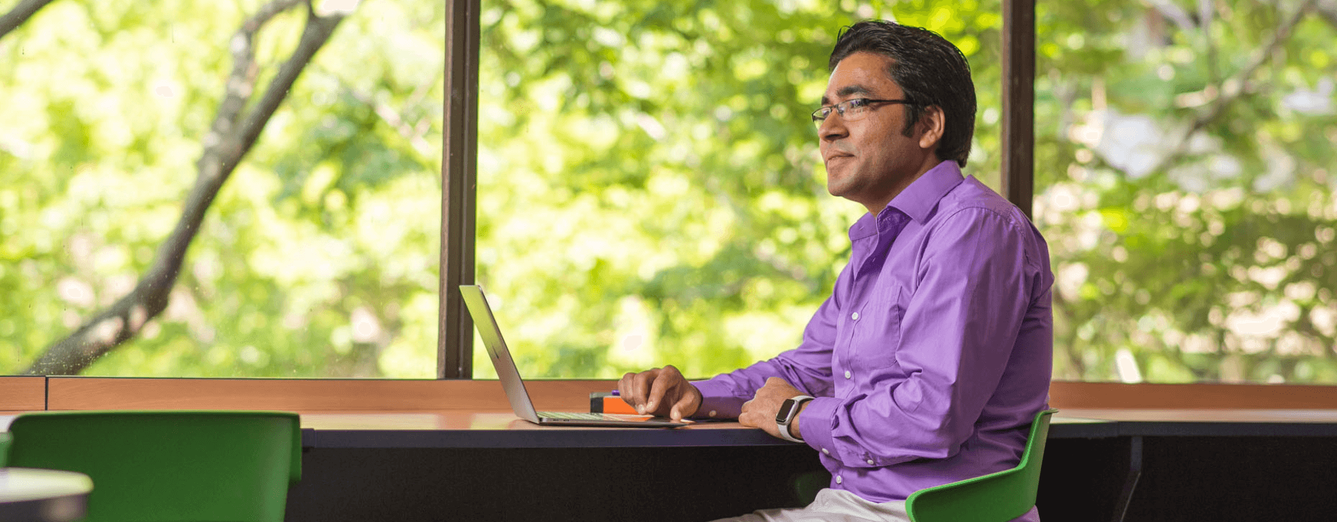 A man in a purple shirt sits at a desk working on a laptop in front of large windows with a view of green trees outside.
