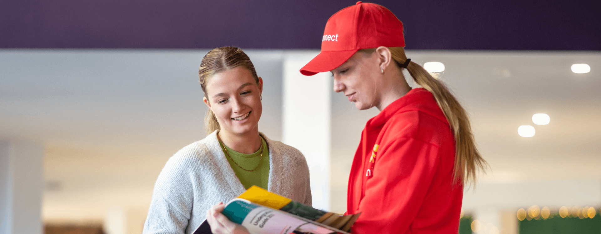 A person in a red uniform and cap shows a magazine or brochure to another person who is smiling.
