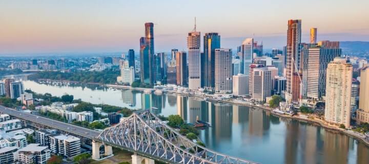 Aerial view of the Brisbane skyline at dusk with a river reflecting the buildings and a steel bridge on the left.