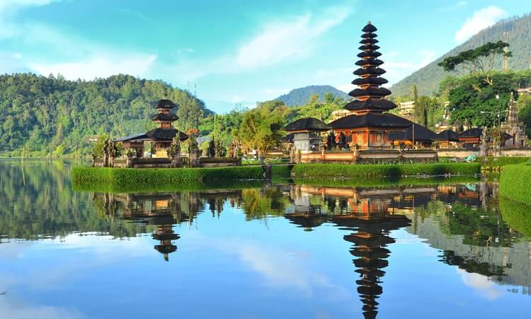 Traditional Balinese temple with multi-tiered pagodas reflected in a serene lake, set against a backdrop of lush greenery and clear blue sky.