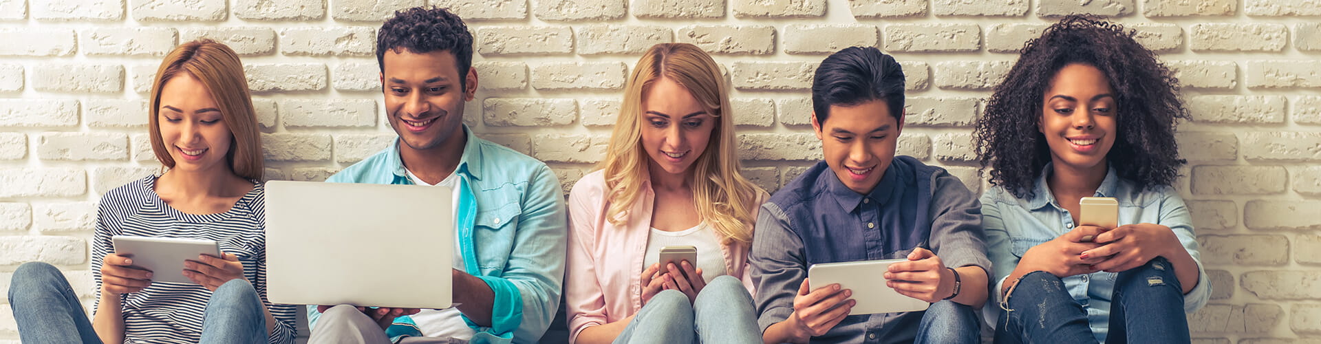 Five individuals seated in a row against a brick wall, each engrossed in their own electronic device, ranging from smartphones to a laptop.