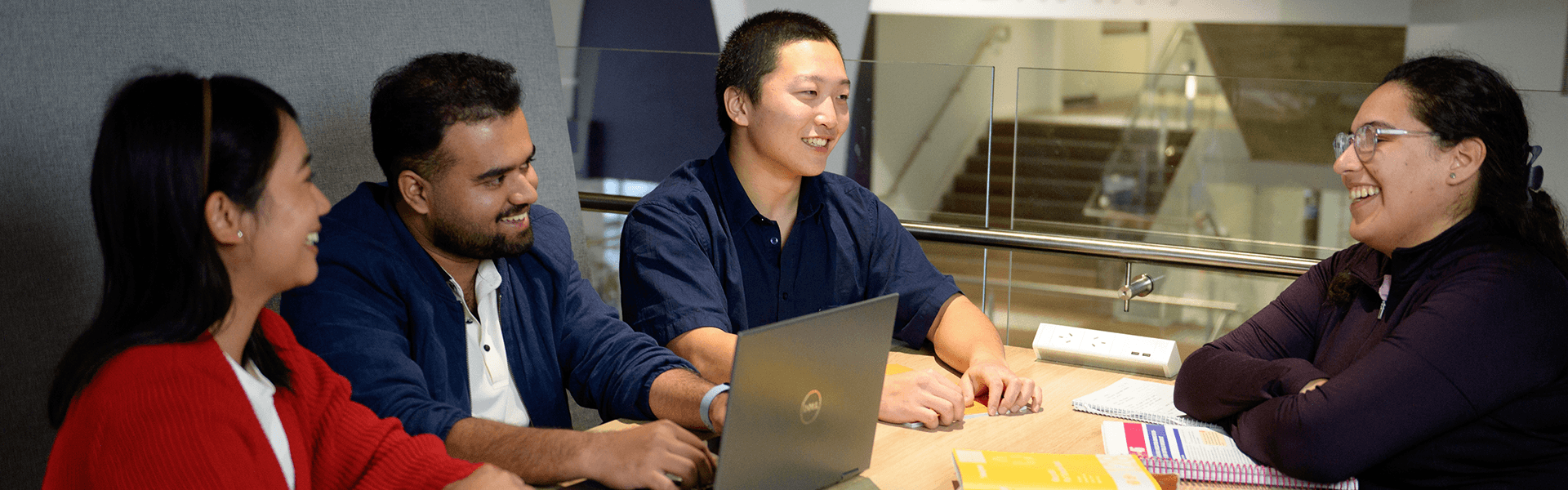 Four people sit around a table with a laptop and books, engaged in conversation and smiling in a modern indoor setting.