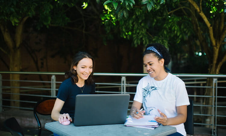 Two individuals studying and working together with a laptop and notebooks at an outdoor table.