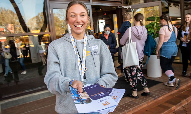 A smiling woman in a hoodie and lanyard handing out pamphlets.