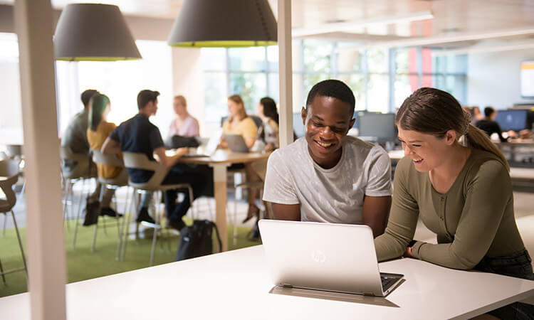 Two people smiling and looking at a laptop in a bright office setting with others in the background.